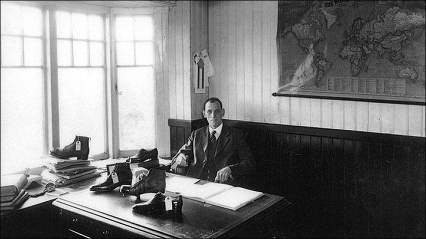 Photograph of Arthur Buckby in his office. taken about  1933, with a selection of boots and shoes on his desk.